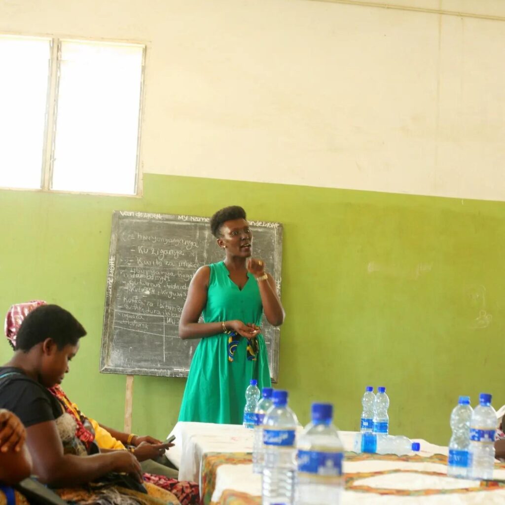A woman in a green dress stands beside a blackboard covered in handwritten notes, speaking to a group seated at a table with water bottles.