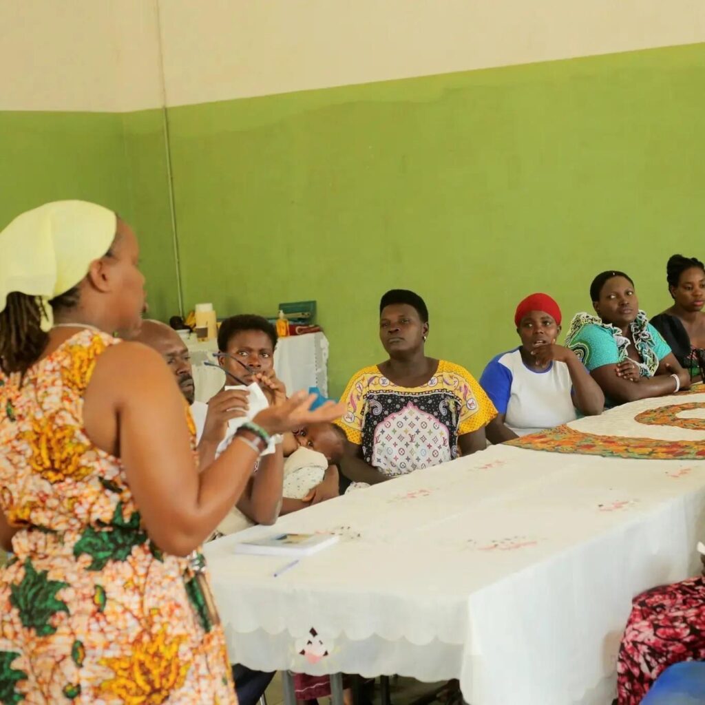 A facilitator in an orange floral dress gestures as she speaks to women seated behind a long table covered with a white embroidered cloth; one participant holds a baby.