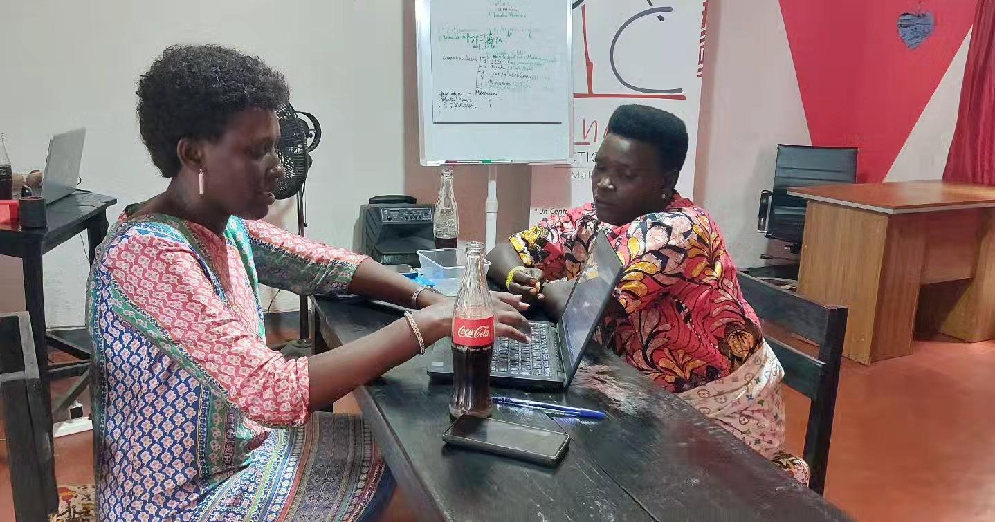 Two women sit at a dark wooden table working on a laptop, with a phone and a bottle of Coca-Cola on the table and an Ingiro Action Center banner and whiteboard behind them.