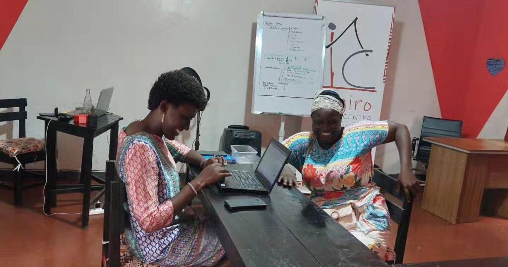 Two women lean over a black laptop at a long table in a red and white room, with a flipchart of handwritten notes and an Ingiro Action Center banner behind them.