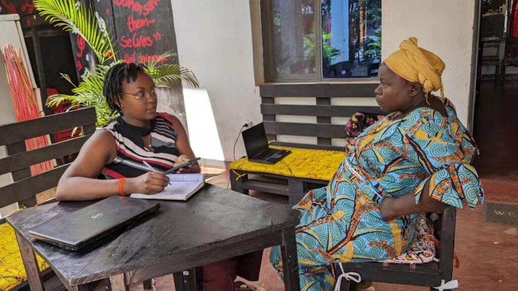 A person wearing glasses holds a pen and an open notebook while talking with a woman in a patterned dress and yellow headwrap seated beside a laptop, with a chalkboard behind them.