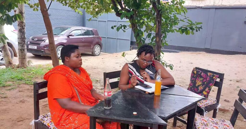 A woman in a bright orange patterned shawl sits at an outdoor table while another person writes in an open notebook, with soft drinks and a phone on the table.