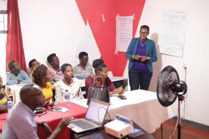A woman in a blue jacket and glasses stands at the front of the room gesturing as she speaks to seated participants, with handwritten flipchart sheets taped to a red and white wall behind her.
