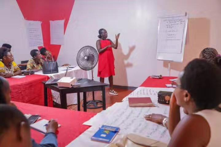 A woman in a red dress stands beside a flipchart on an easel speaking to participants seated at tables covered with red cloths, notebooks and pens in front of them.