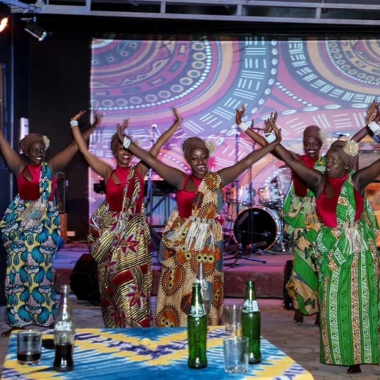 Dancers in red tops and matching patterned skirts perform on stage with their arms raised, in front of a colourful circular backdrop and a drum kit.