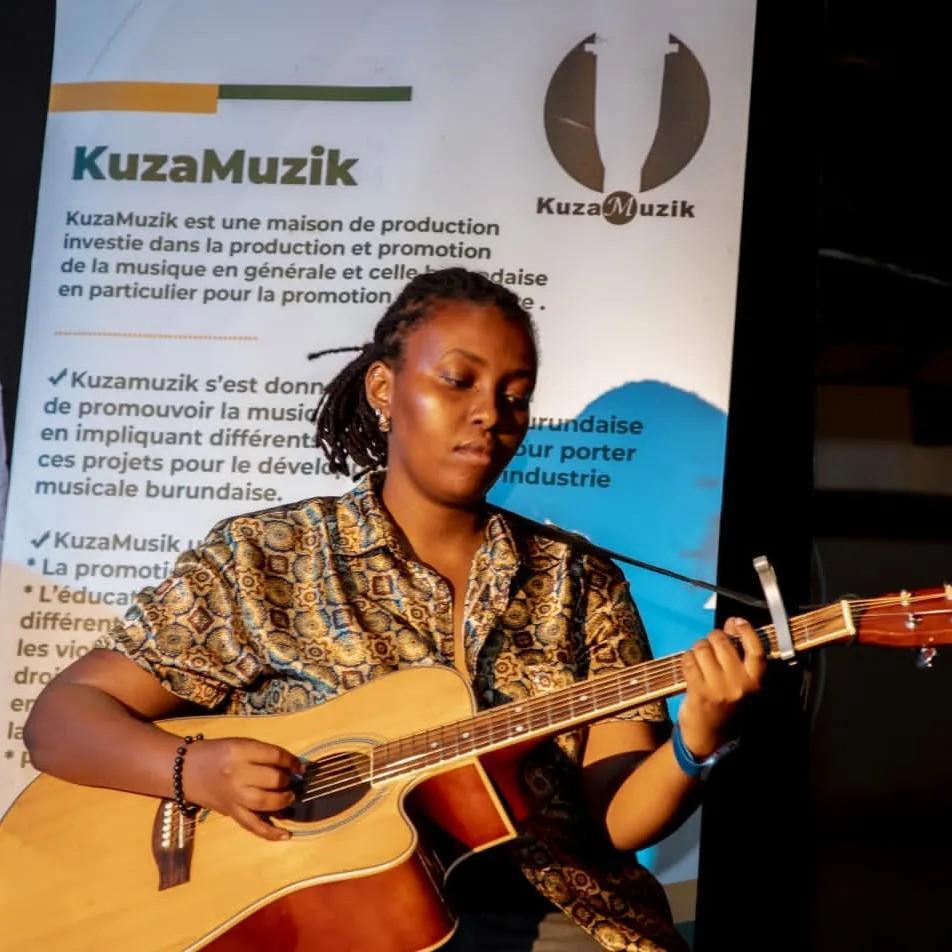 A woman with braided hair plays an acoustic guitar on stage in front of a KuzaMuzik banner.