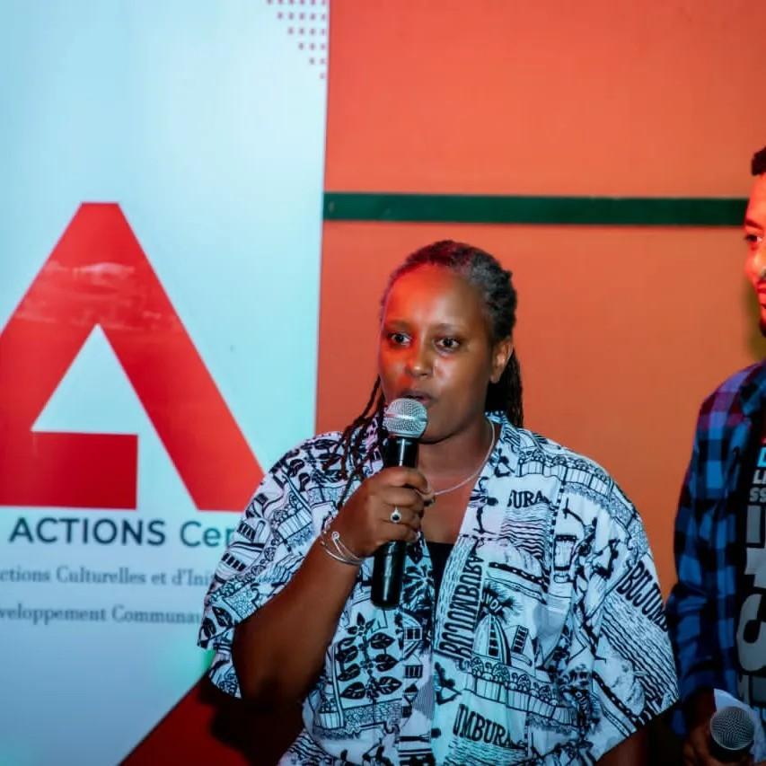 A woman with braided hair speaks into a microphone in front of an Ingiro Actions Center banner, with a second presenter holding a microphone beside her.