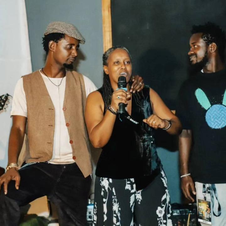A woman holds a microphone while standing between two people on a dimly lit stage.