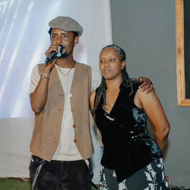 A person wearing a light vest and cap holds a microphone beside another person on stage.