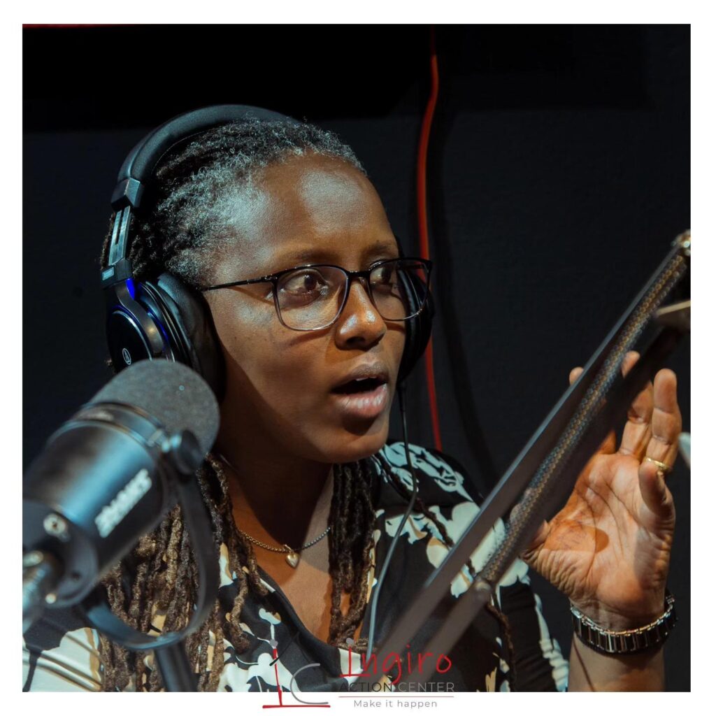 A woman wearing glasses and headphones speaks into a microphone and raises one hand.
