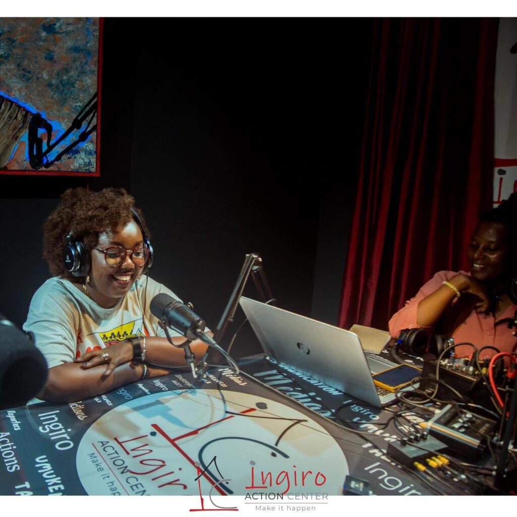 Two people sit at a studio table with a laptop, microphone, and audio equipment.