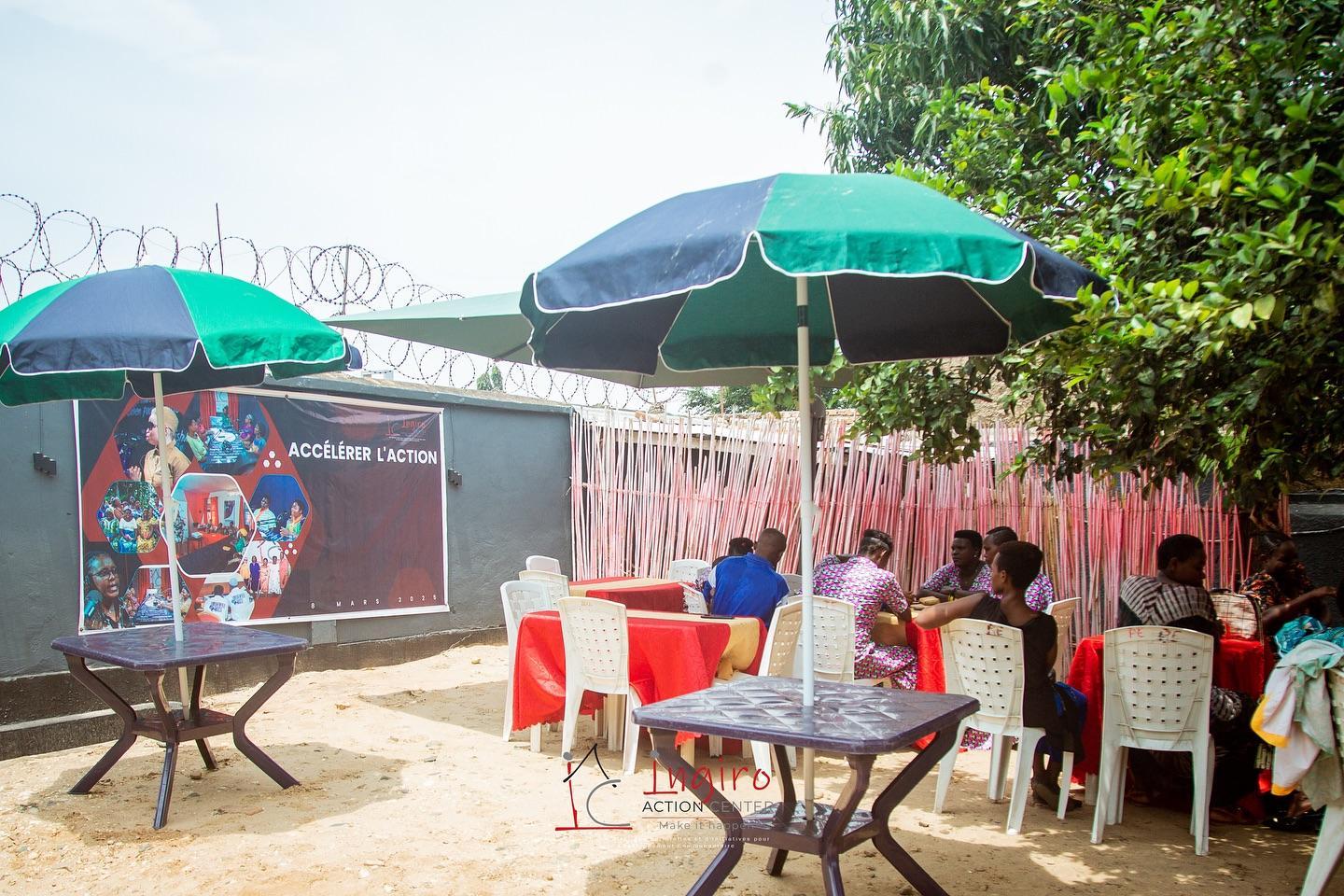 People sit at tables in an outdoor gathering space decorated with red fabric and greenery.