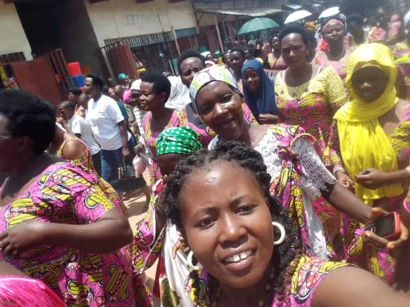 A smiling woman in a bright patterned outfit stands among a crowd of people.
