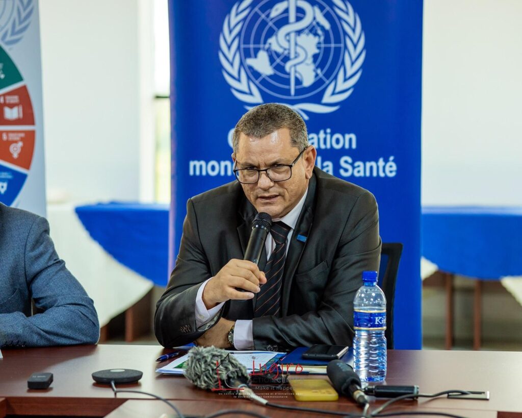 A man in a dark suit sits at a conference table beneath a blue United Nations emblem.