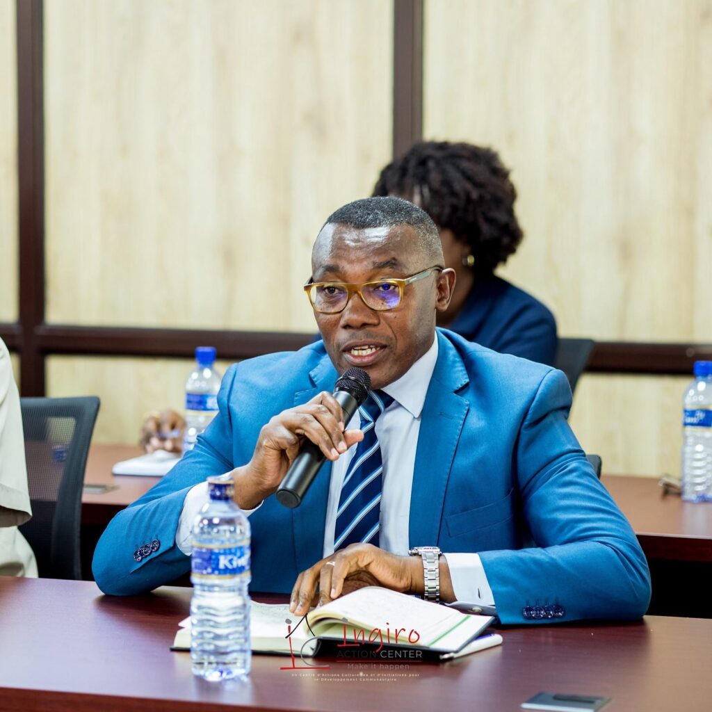 A man in a blue suit sits at a conference table with a microphone and a bottle of water.