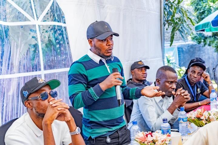 A man in a cap and striped shirt holds a microphone near a seated group.