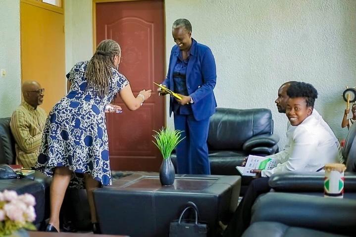 A woman in a blue-patterned dress reaches toward a yellow item held by a person in a blue suit while others watch.