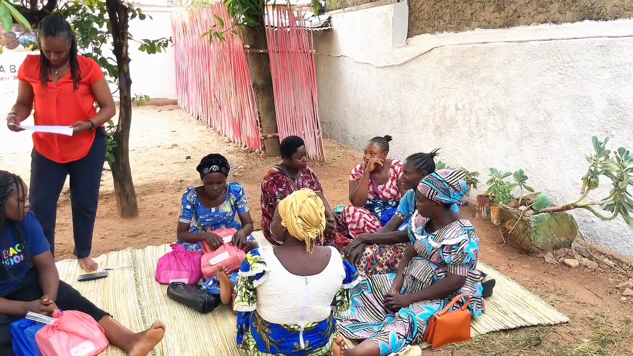 Several children sit outdoors with colorful bags and books in front of them; an adult in a red top bends toward the group.