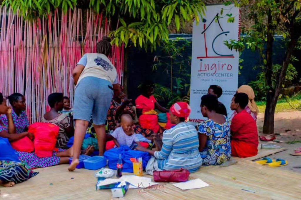 Children and adults sit together on the floor in a decorated room with bags and books nearby.