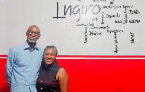 A man in a light blue shirt and a woman in a dark sleeveless top stand together in front of a red wall with painted words.