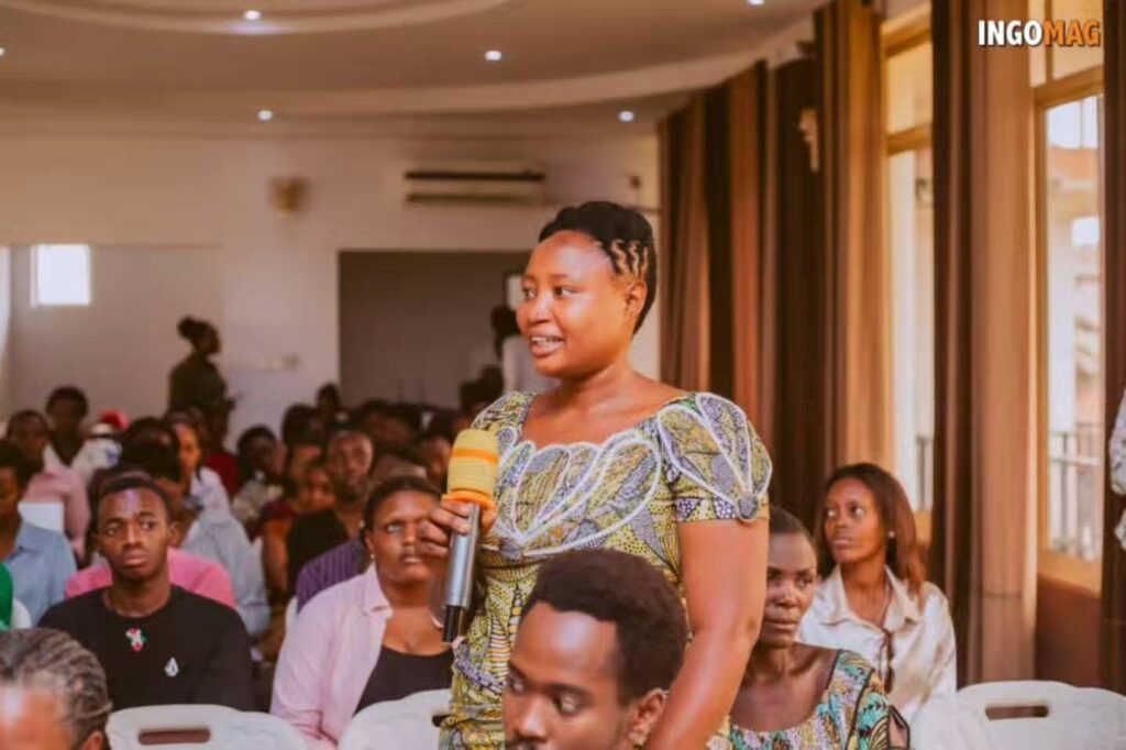A woman in a patterned dress stands holding a microphone in front of seated attendees.