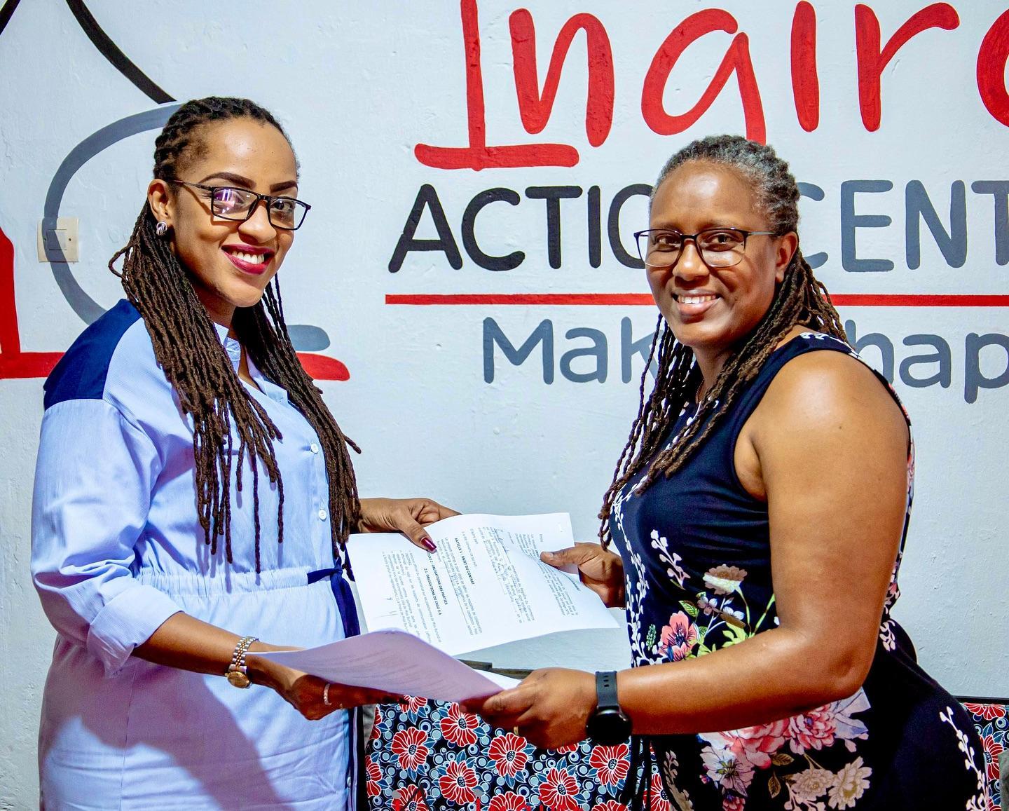 Two women stand side by side holding documents in front of an Ingiro Action Center sign.