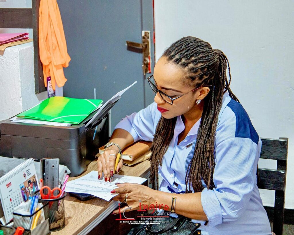 A woman in a blue shirt writes on a document at a desk while another person sits nearby.