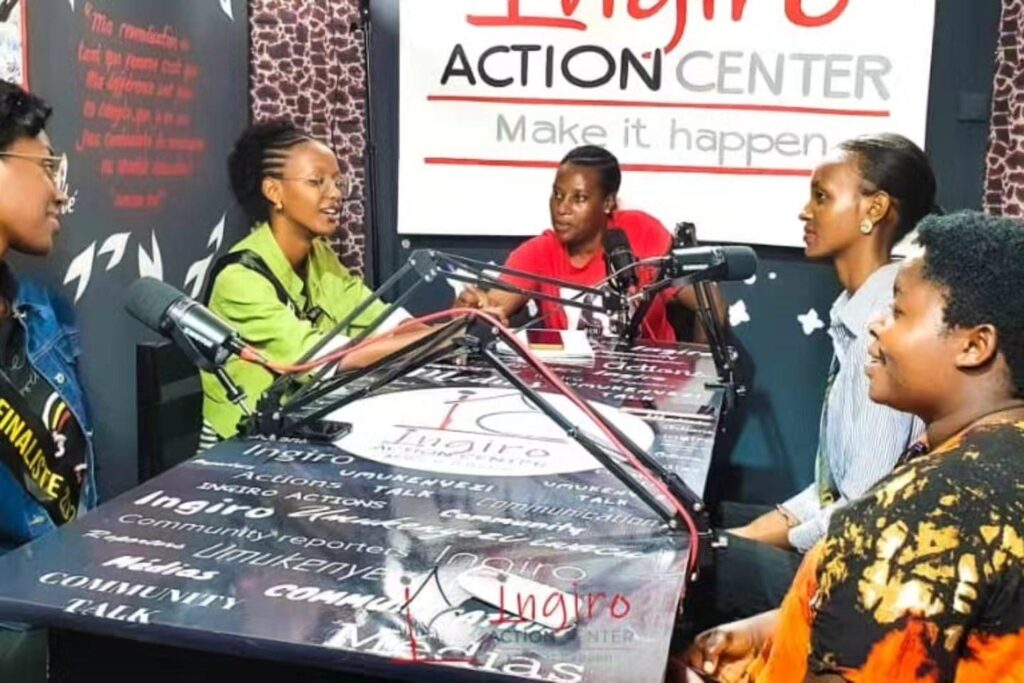 Two women sit at a studio table with a microphone and a patterned backdrop.