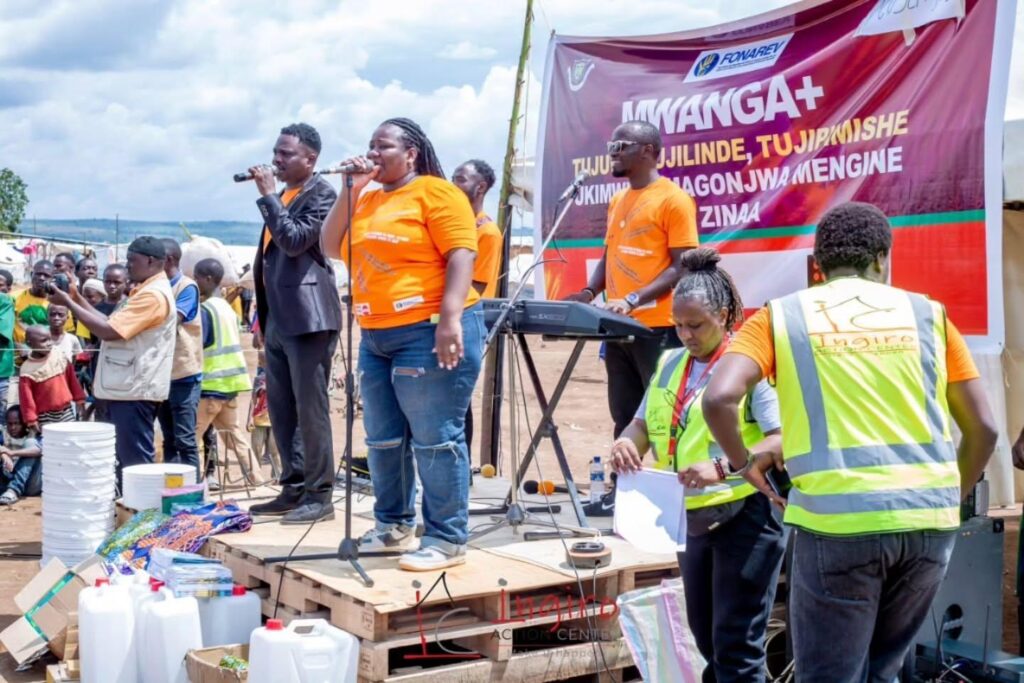 People stand near a stage and microphone beneath a Mwanga+ banner.