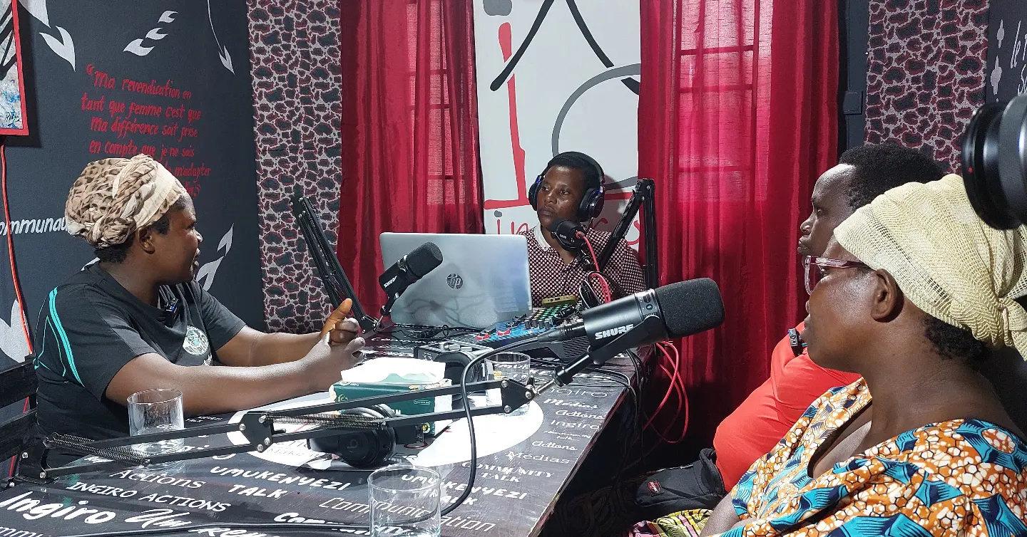 People sit around a table in a radio studio with microphones and recording equipment.