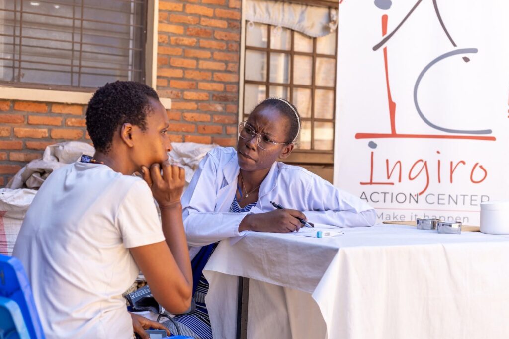 A woman sits across a table from a health professional in a white coat who is writing notes, beside an Ingiro Action Center banner.