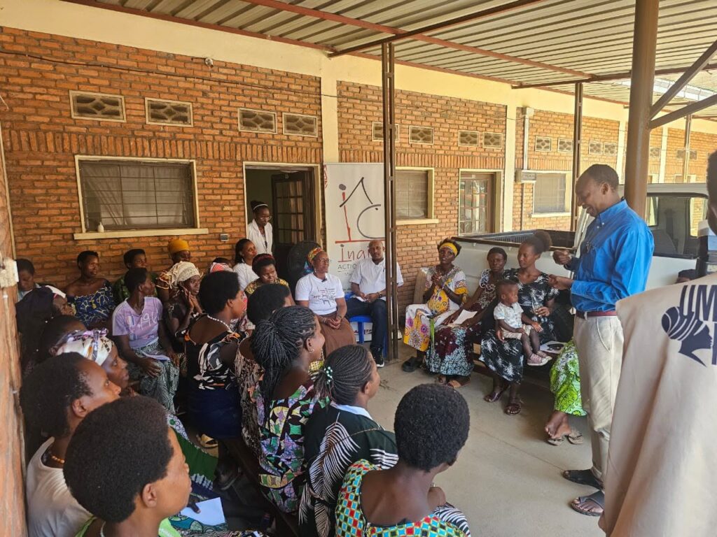 A man in a blue shirt speaks to women seated closely together under a covered walkway.