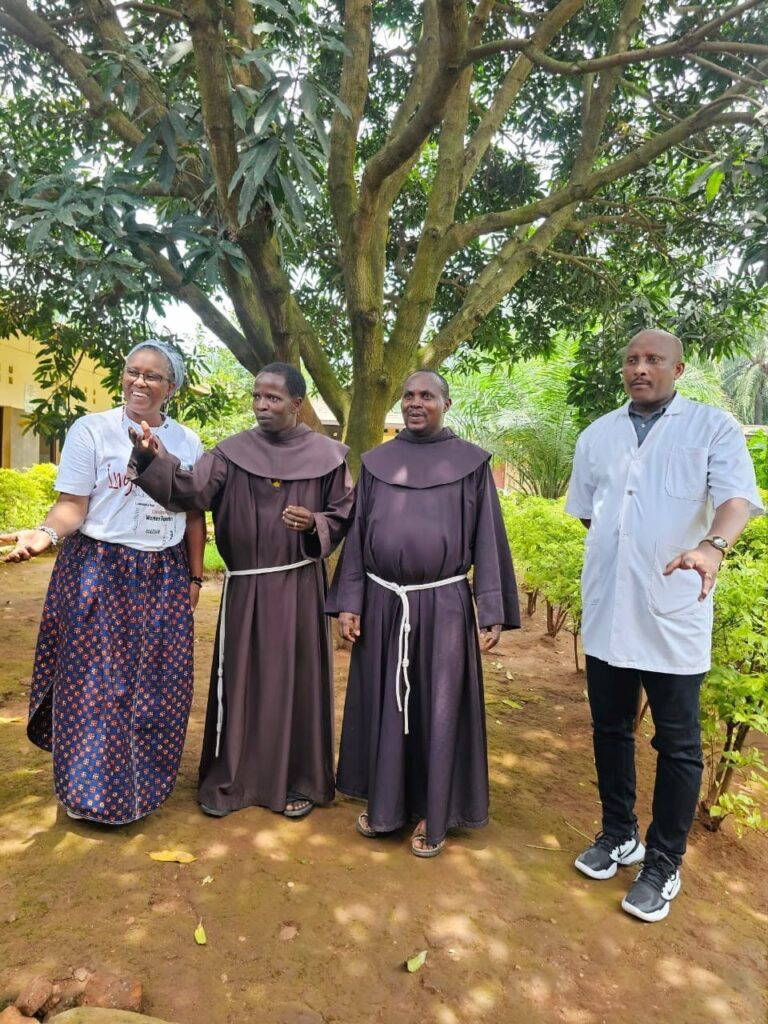 Two parish priests in brown habits stand with a woman in a patterned skirt and a man in a white coat under a mango tree.
