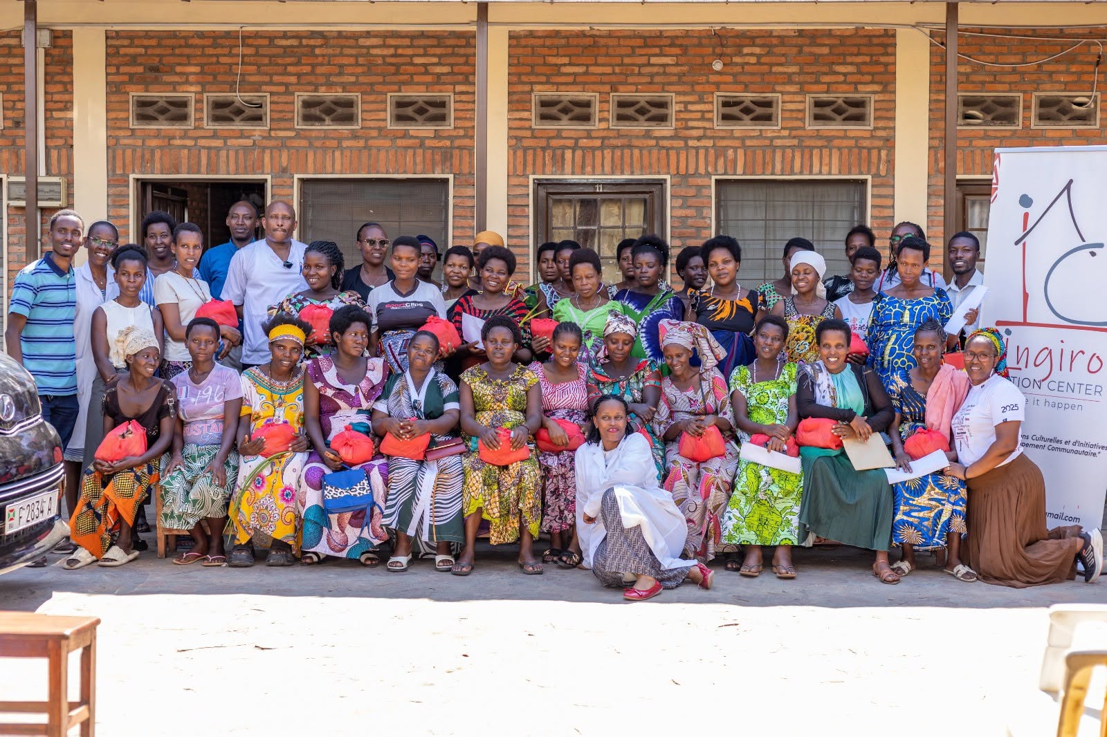 Women and members of the team pose for a group photograph in front of a brick building and an Ingiro Action Center banner.