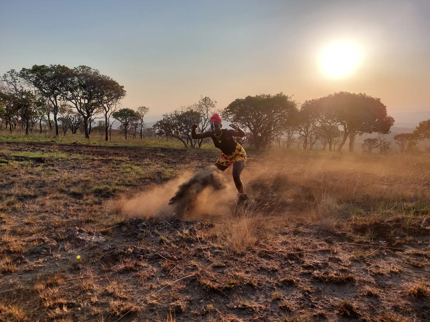 A dancer kicks up dust against a setting sun, with trees on a dry plain.