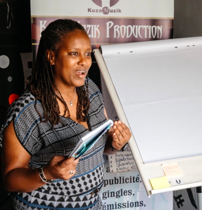A woman speaks beside a flipchart in front of a Kuza Muzik Production banner.