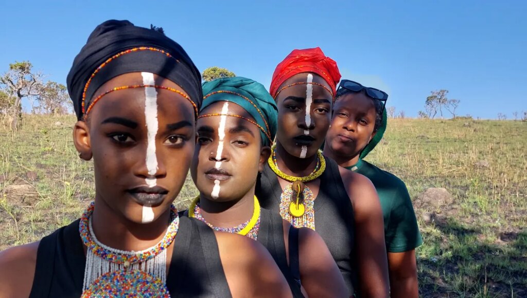 Four people with white face paint, beaded necklaces and head wraps pose outdoors.