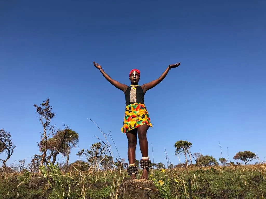 A dancer in a colourful skirt stands with arms outstretched under a blue sky.