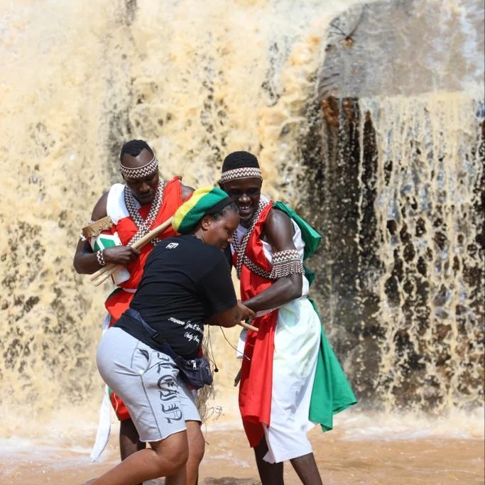 Two performers in red, white and green garments dance in front of a waterfall.