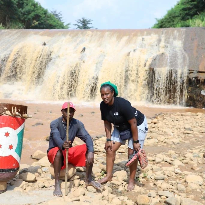 A man seated on rocks beside an object painted in the colours of the Burundian flag, and a woman in a black T-shirt, in front of a waterfall.