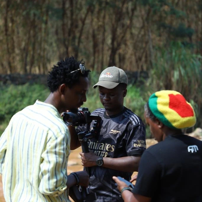 A cameraman films while a woman in a red, yellow and green hat and a man in a striped shirt look on.